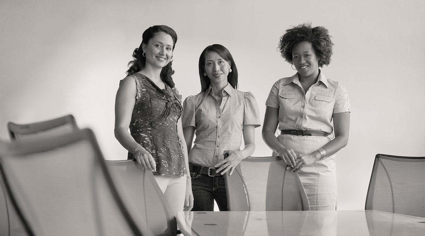 Three women standing behind the chairs of a boardroom table, smiling at the camera