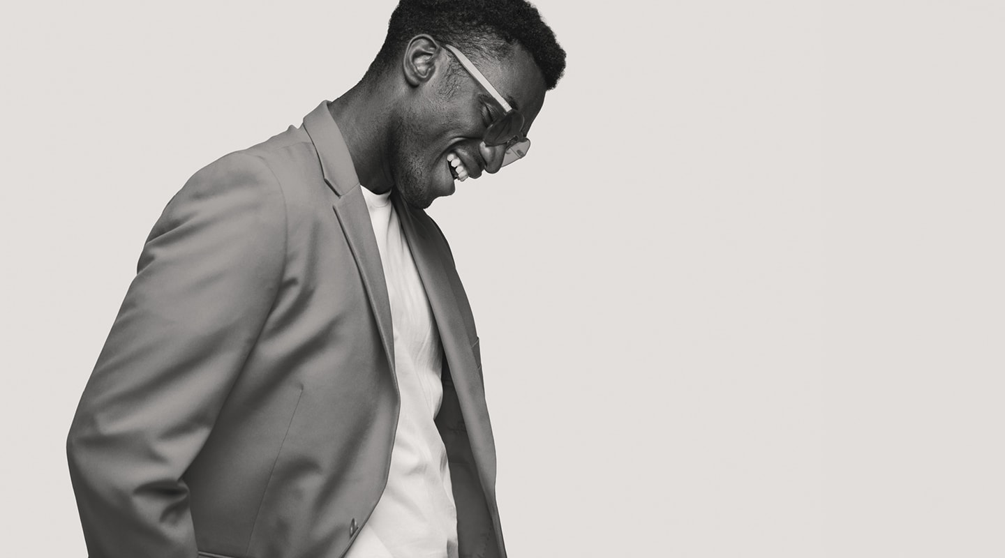 A smiling man in a light suit and sunglasses, looking down