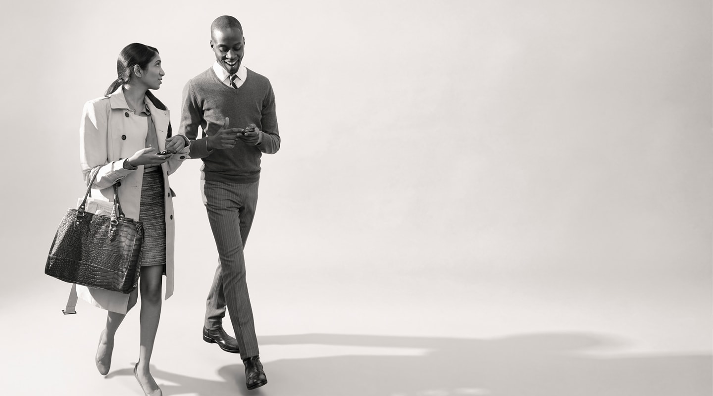 A woman with a handbag and a man in a sweater and tie walking and talking, phones in hand