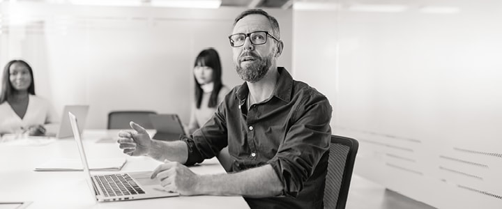 A man in glasses making a point at a meeting table, two colleagues listening behind him