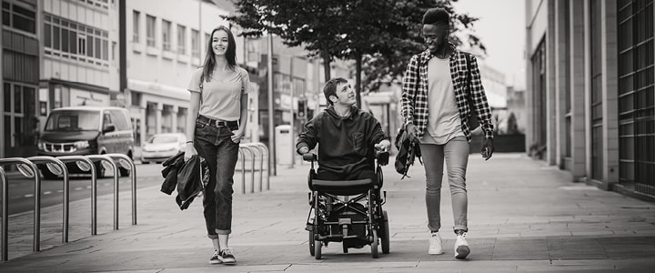 Three young friends, one in a power wheelchair, walking and talking along a city street