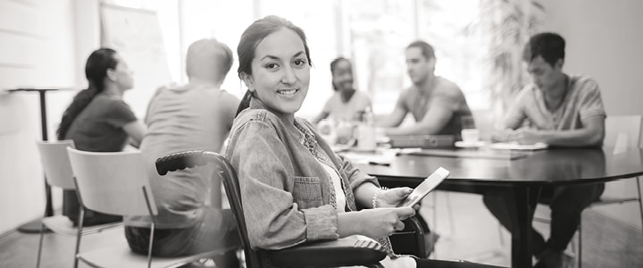 A smiling young woman in a wheelchair holding a tablet, a group working at a table behind her