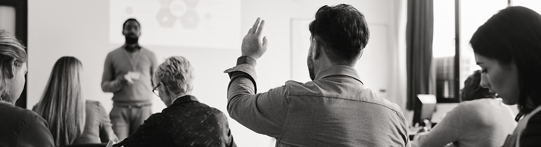 A student raising a hand in a lecture room while a presenter speaks at the front