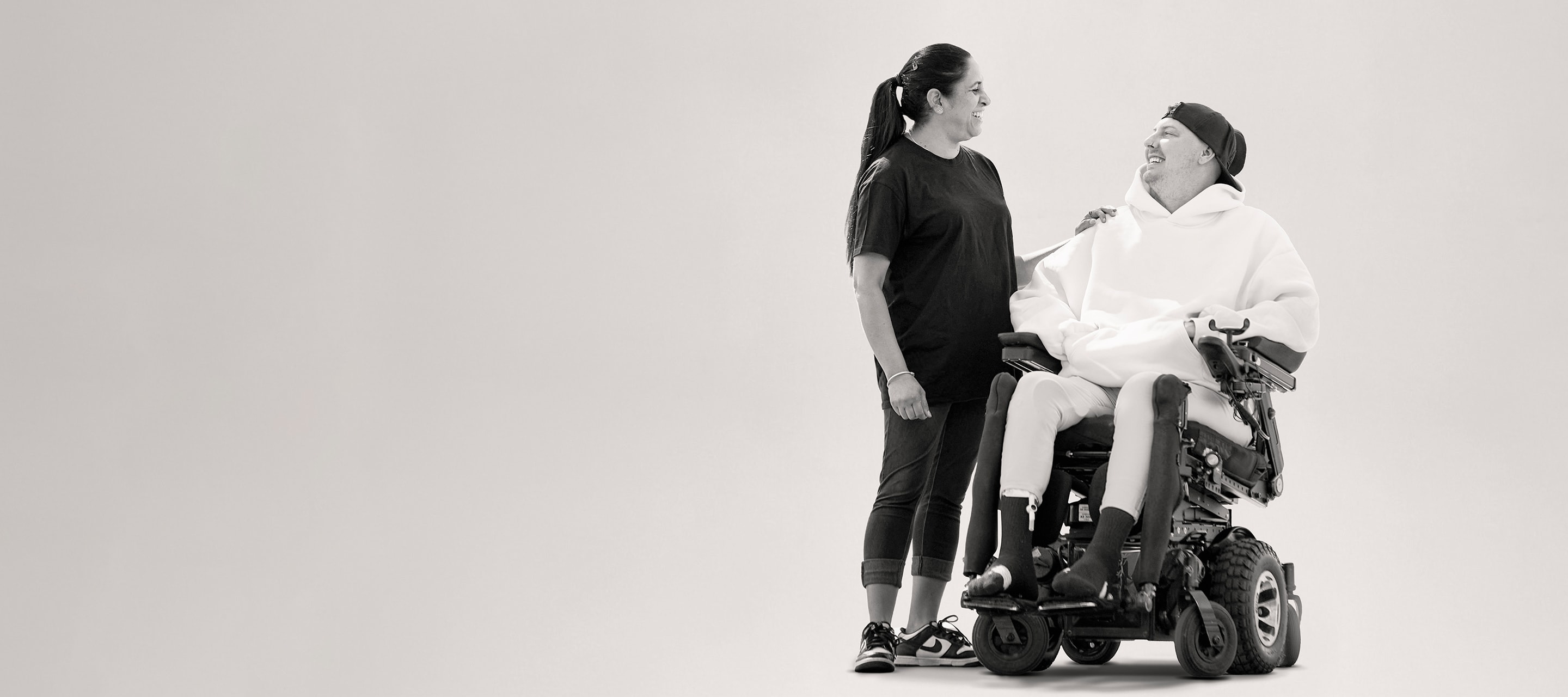 A carer with her hand on the shoulder of a smiling young man in a power wheelchair